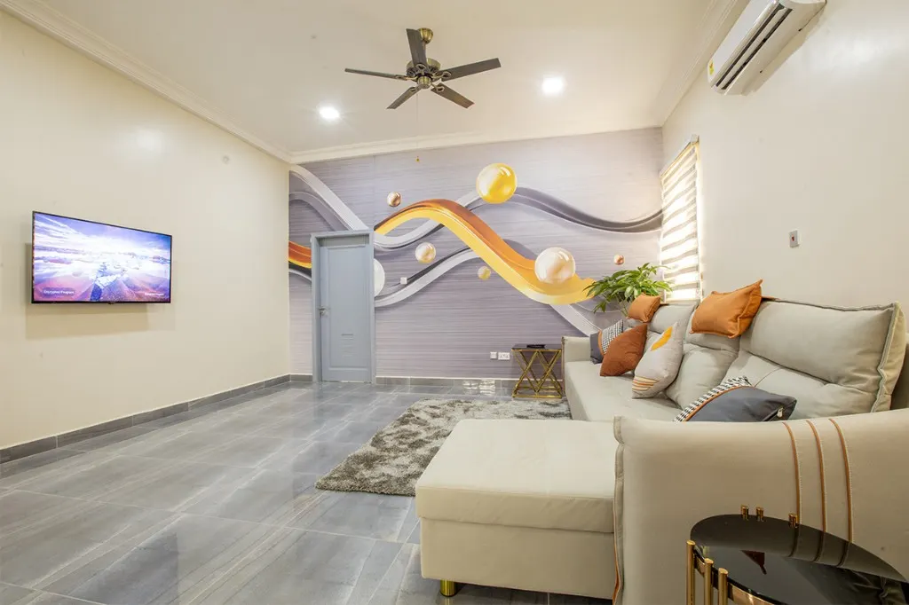 Lounge at Plantsville in Dansoman, Accra, with a grey sofa, orange cushions, a wall-mounted television, and an abstract mural