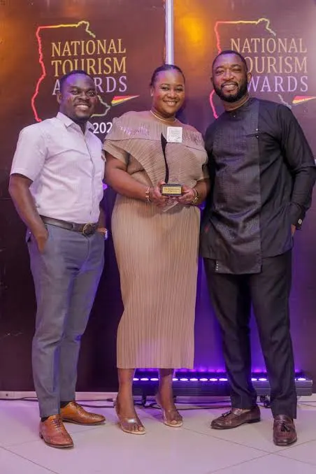 Three people standing together, a woman holding an award, with National Tourism Awards logos behind them